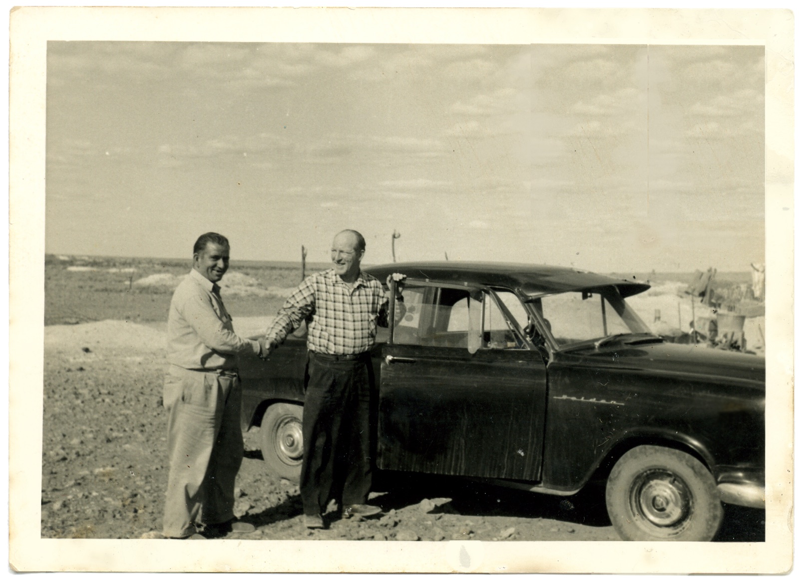 George Christianos Sr shakes hands with a French opal buyer beside a Holden on the Coober Pedy opal fields, 1960s